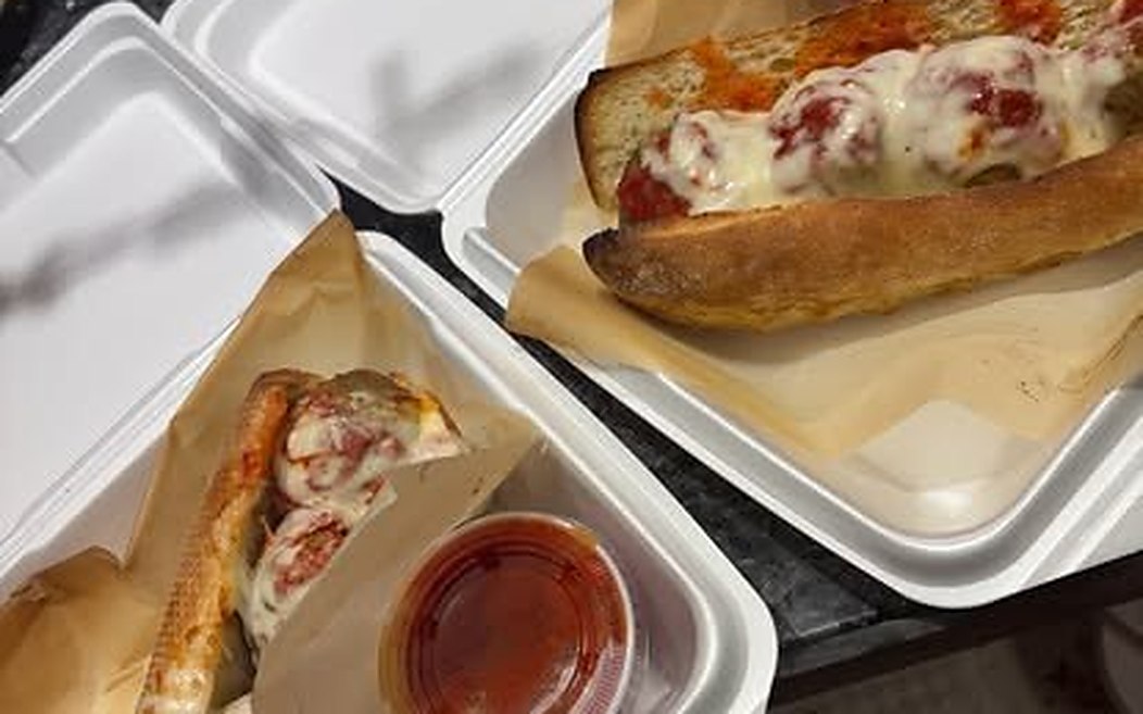 A toasted meatball sub and a pizza slice packed for carryout with sauce cups